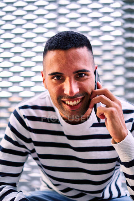 Feliz hombre hispano en ropa casual mirando a la cámara con sonrisa y hablando en el teléfono inteligente mientras está sentado en la frontera contra la pared ornamental en la calle de la ciudad - foto de stock