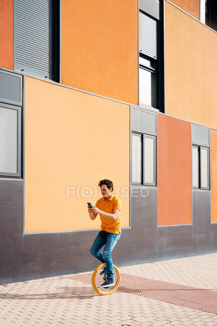 Corps complet de jeune homme agile utilisant le téléphone mobile tout en chevauchant monocycle près de bâtiment urbain coloré moderne — Photo de stock