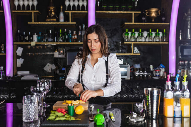 Young female barkeeper in stylish outfit preparing mojito cocktail with lemon slices while standing at counter in modern bar — Stock Photo