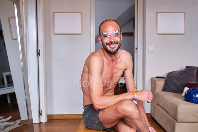 Vue de face de gai homme barbu torse nu avec drag queen maquillage souriant et regardant la caméra tout en étant assis près du miroir à la maison — Photo de stock