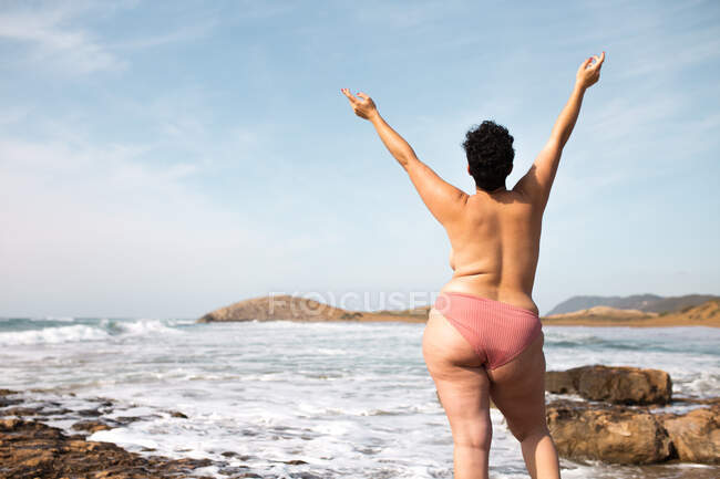 Rückenansicht einer gesichtslosen Oben-ohne-Frau in Unterhosen, die mit erhobenen Händen an der felsigen Küste mit Felsbrocken in der Nähe des Meeres bei Tag unter dem Himmel steht — Stockfoto