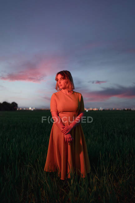 Verträumte junge Frau im Vintage-Kleid schaut nachdenklich weg, während sie allein im dunklen Grasfeld vor wolkenverhangenem Abendhimmel in der Dämmerung steht — Stockfoto
