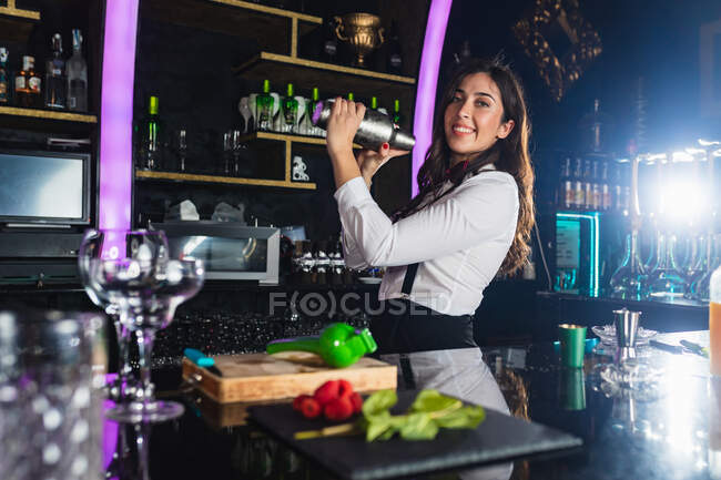Счастливая барменша в стильном наряде, трясущая металлическим шейкером, пока готовит коктейль стоя у стойки в современном баре — стоковое фото