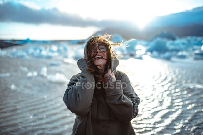 Молодий сміється турист в окулярах з пірсингом і вітряним волоссям біля води в сонячний день на розмитому фоні — стокове фото