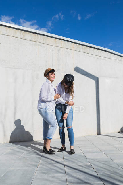 Full body of young positive female friends in trendy outfits and hats standing on walkway near gray wall in sunny day under blue sky — Stock Photo