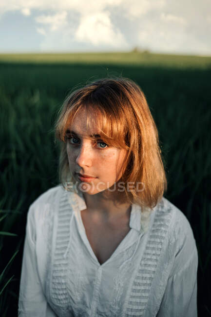 Jeune femme paisible en chemisier blanc de style rétro assis au milieu de hautes herbes vertes et regardant la caméra tout en se reposant dans la soirée d'été dans la campagne — Photo de stock