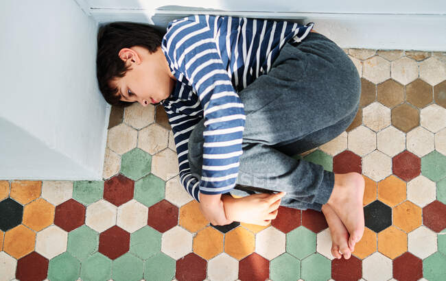 Desde arriba de infeliz niño preadolescente solitario acostado en el suelo y abrazando las rodillas como víctima de violencia doméstica y abuso - foto de stock