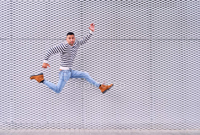 Pieno corpo eccitato maschio ispanico sorridente e saltando sul marciapiede contro muro grigio ornamentale divertendosi sulla strada della città — Foto stock