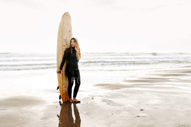 Mujer surfista vestida con traje de neopreno de pie mirando hacia otro lado con la tabla de surf en la playa durante el amanecer en el fondo - foto de stock
