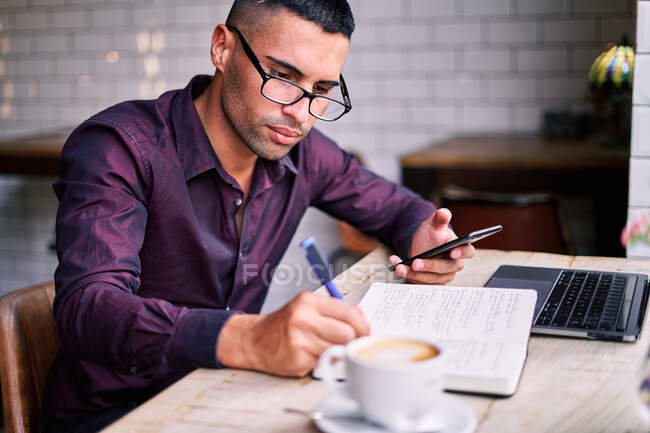 Seitenansicht eines hübschen hispanischen Jungunternehmers, der Informationen auf dem Smartphone überprüft und Notizen im Planer schreibt, während er am Tisch mit Laptop arbeitet — Stockfoto