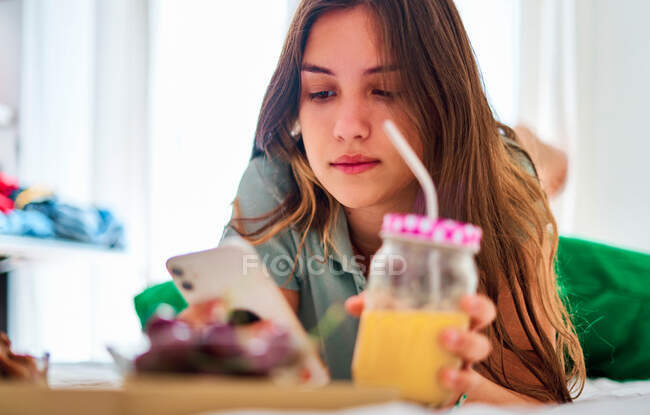 Joven estudiante que navega por las redes sociales en el teléfono móvil cerca de la mesa con frutas frescas y jugo mientras pasa la mañana en casa - foto de stock