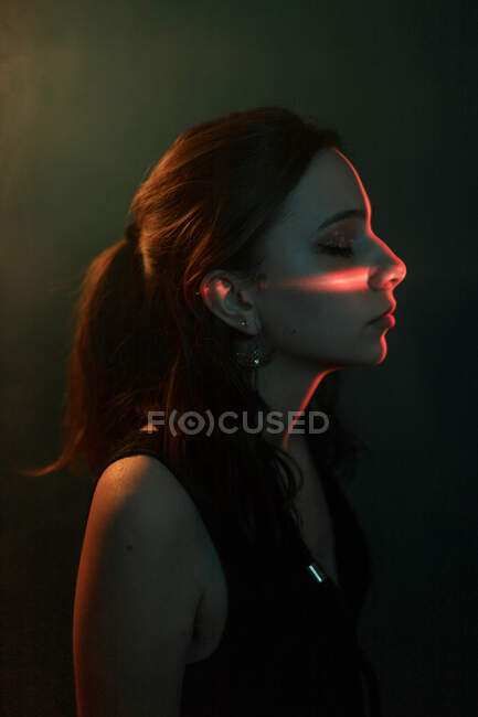 Vue latérale du jeune modèle féminin avec projection de lumière sur le visage debout dans un studio sombre avec les yeux fermés — Photo de stock
