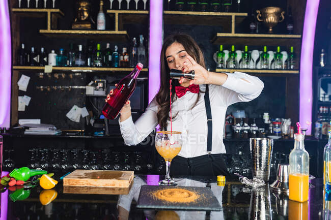 Сфокусований жіночий бармен у стильному вбранні, що додає рідину з пляшки в джиґер, готуючи коктейль, що стоїть за стійкою в сучасному барі — стокове фото