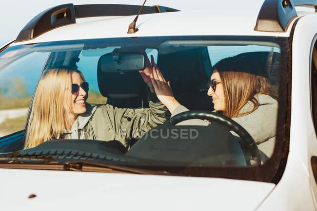 Веселі друзі-жінки сидять в машині і дарують високі п'ять під час подорожі на сонячну природу — стокове фото
