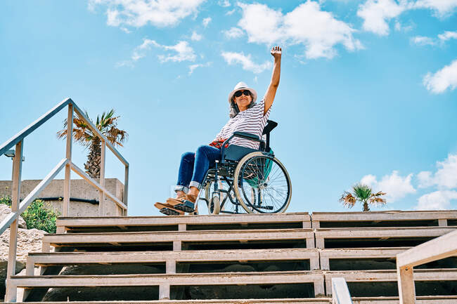 Niedriger Winkel Ganzkörper positiv behinderter Frau sitzt im Rollstuhl in der Nähe der Treppe und winkt mit der Hand gegen den blauen Himmel in tropischen Stadt — Stockfoto