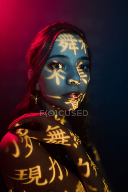 Модная молодая женская модель со светлой проекцией в виде восточных иероглифов, смотрящая на камеру в темной студии с красной подсветкой — стоковое фото