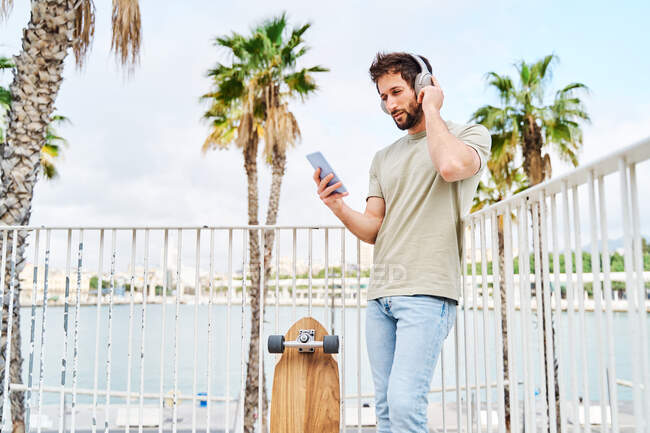 Full body of male in wireless headphones leaning on fence and using mobile phone standing near skateboard — Stock Photo