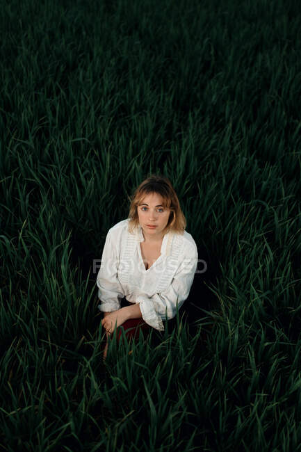 Mujer joven pacífica en blusa blanca de estilo retro sentada en medio de una hierba verde alta y mirando a la cámara mientras descansa en la noche de verano en el campo - foto de stock