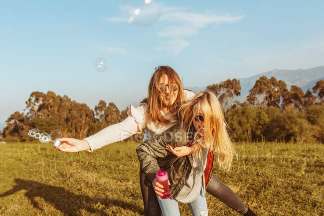 Rindo mulher loira dando passeio de piggyback para namoradas alegres segurando sabão bolha vara andando na natureza — Fotografia de Stock