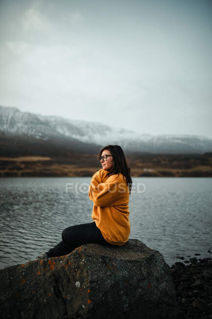 Vista laterale del giovane turista premuroso in occhiali da vista seduto sulla pietra sulla costa e guardando lontano vicino alla superficie dell'acqua e montagne — Foto stock