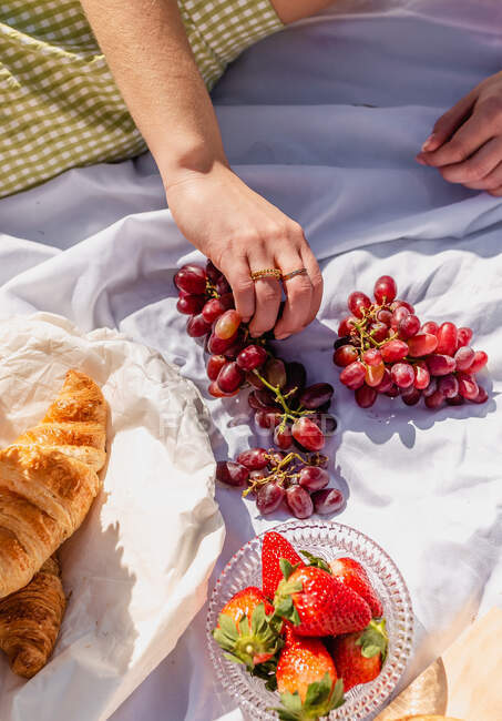 High angle of crop female with ripe grapes having picnic with croissant and strawberries with focaccia — Stock Photo