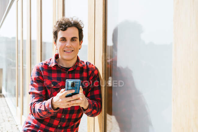 Jeune homme en tenue décontractée utilisant un smartphone tout en regardant la caméra contre le bâtiment urbain moderne — Photo de stock