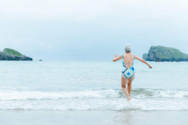 Back view active fit aged female in swimsuit walking on sea water while enjoying summer day on beach — Stock Photo