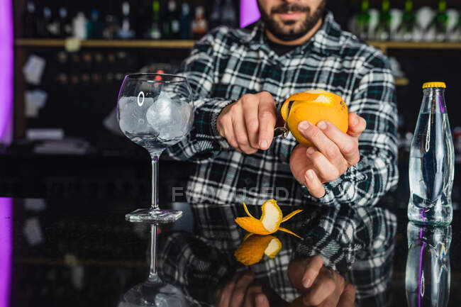 Cortado irreconocible camarero masculino añadir cáscara de naranja en un vaso mientras se prepara cóctel de pie en el mostrador en el bar moderno - foto de stock