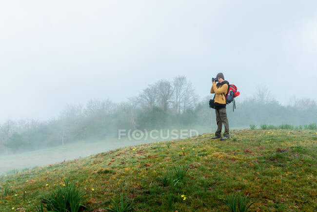 Seitenansicht einer Backpackerin, die in einer nebligen Wiese steht und während der Fahrt die bergige Landschaft fotografiert — Stockfoto