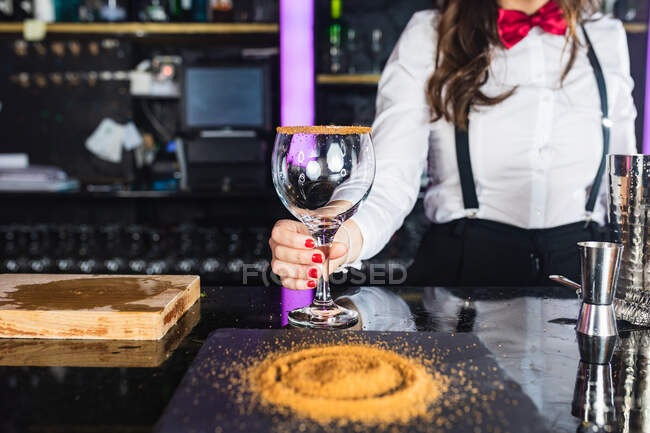 Cropped unrecognizable female barkeeper in stylish outfit preparing cocktail with sugar on glass while standing at counter in modern bar — Stock Photo