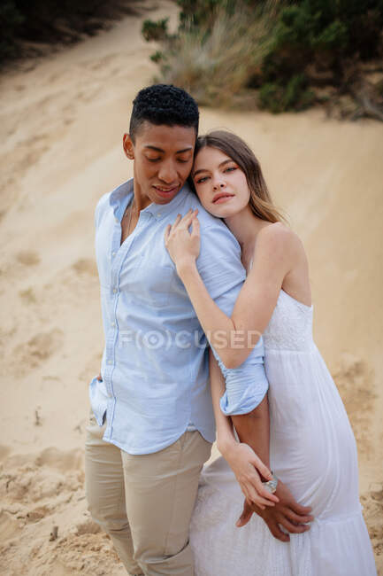 Side view of loving multiracial couple of newlyweds hugging while standing on sandy hill on wedding day — Stock Photo