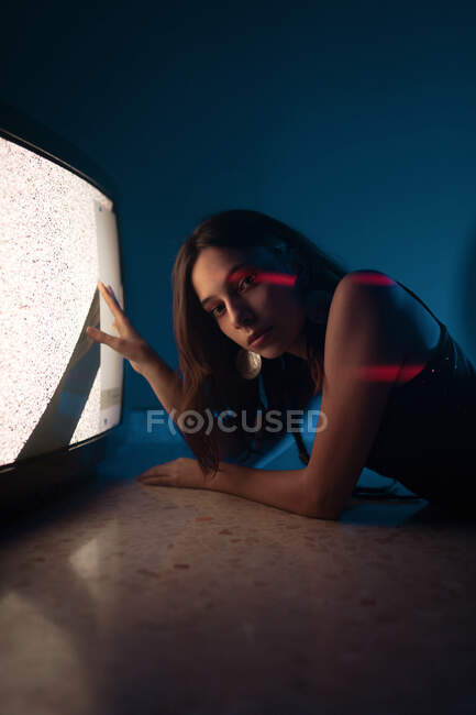 Side view of female model in black dress lying down on floor looking at camera touching glowing old television in dark studio — Stock Photo