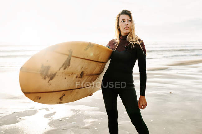 Surfer donna vestita in muta in piedi guardando lontano con la tavola da surf sulla spiaggia durante l'alba sullo sfondo — Foto stock