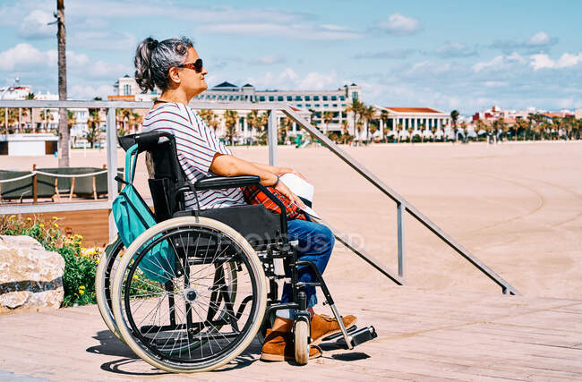 Вид збоку жінка мандрівник в інвалідному візку з рюкзаком насолоджується літньою подорожжю на пляжі біля блакитного моря — стокове фото