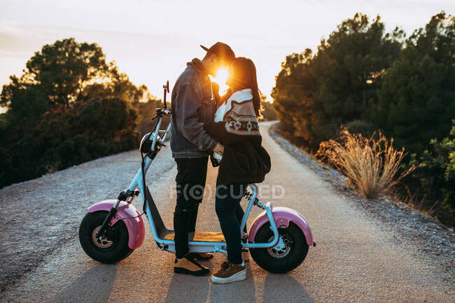 Vista lateral de los jóvenes hipsters abrazando cerca de scooter vintage en la ruta del campo al atardecer - foto de stock