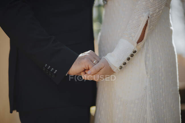 Vista lateral da colheita do noivo anônimo segurando as mãos de noiva elegante em vestido de casamento branco — Fotografia de Stock