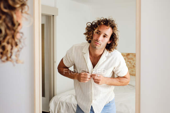 Reflexão de macho elegante com cabelo encaracolado abotoando camisa enquanto olha no espelho no quarto — Fotografia de Stock