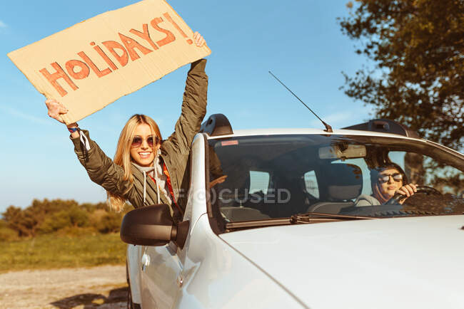 Mujer alegre que sobresale por la ventana del coche mostrando cartón con vacaciones mientras conduce en coche con su novia en la naturaleza - foto de stock