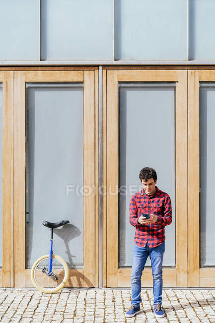 Повний набір молодих чоловіків у повсякденному одязі з використанням смартфона, стоячи біля припаркованого одноколісного велосипеда проти сучасного міського будинку. — стокове фото