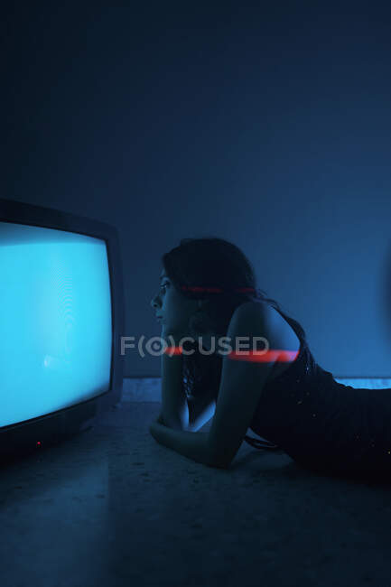 Vista lateral do modelo feminino em vestido preto deitado no chão perto de uma televisão antiga brilhante no estúdio escuro — Fotografia de Stock