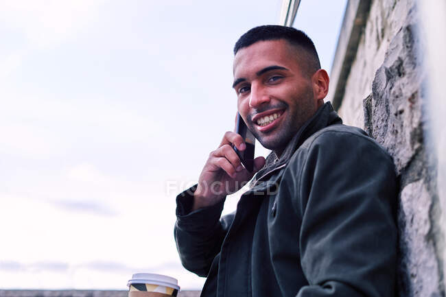 Low angle of glad Hispanic man with coffee to go smiling and looking at camera while leaning on stone wall and speaking on cellphone on street — Stock Photo