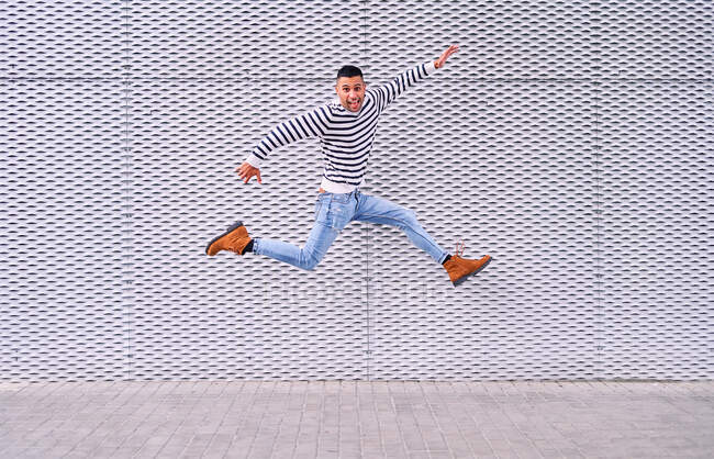 Ganzkörper aufgeregten hispanischen Mann lächelt und springt über Bürgersteig gegen ornamentale graue Wand, während Spaß auf der Straße der Stadt — Stockfoto