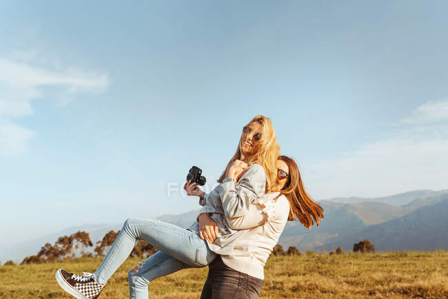 Cheerful woman carrying happy girlfriends in arms spending time together on field in high mountains in sunlight — Stock Photo