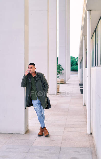 Full body Hispanic man in casual clothes looking away and speaking on mobile phone while standing on pavement before work — Stock Photo