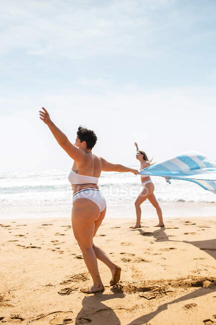 Visão lateral de corpo inteiro de amigas anônimas em trajes de banho passeando na costa arenosa com toalha perto do oceano sob o céu azul nublado em dia ensolarado — Fotografia de Stock