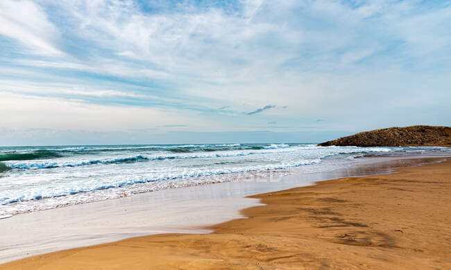 Vista mozzafiato dell'oceano ondulato schiumoso vicino alla costa sabbiosa situata sotto il cielo nuvoloso e luminoso alla luce del giorno — Foto stock