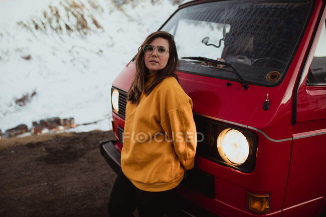 Jeune touriste à lunettes debout près de l'automobile vintage entre terrain désert dans la neige près des montagnes — Photo de stock