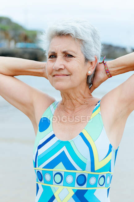 Happy aged gray haired female in stylish bright swimwear keeping hands behind head while doing exercises on beach near sea — Stock Photo
