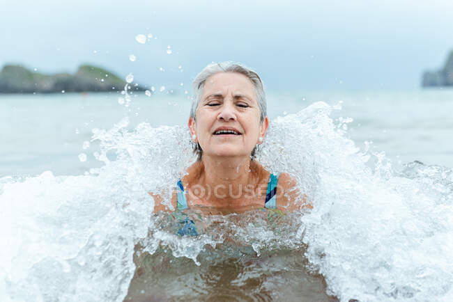 Mujer de pelo gris senior activa nadando en el agua del océano mientras disfruta del verano y practica un estilo de vida saludable en la orilla del mar - foto de stock