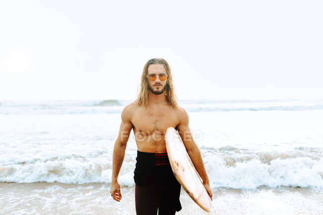 Young surfer man with long hair dressed in wetsuit and stylish sunglasses standing looking at camera with surfboard towards the water to catch a wave on the beach — Stock Photo